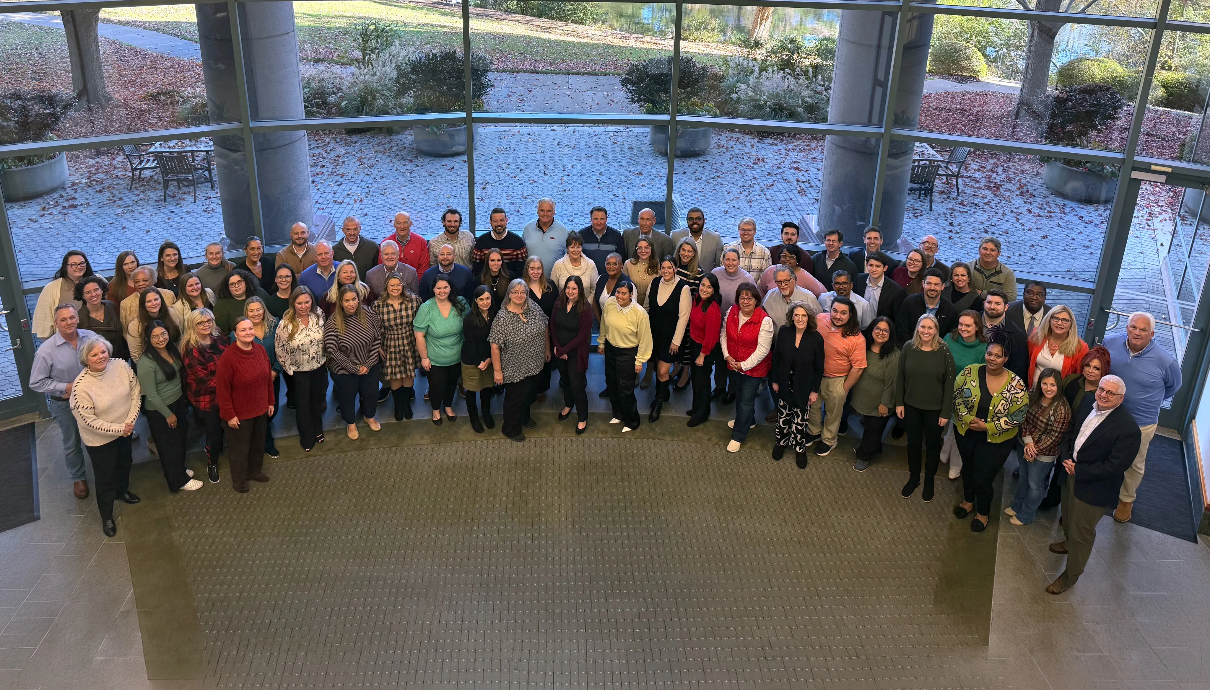 Large group of diverse people posing inside a building with large glass windows showing an outdoor garden area with trees and fallen leaves.