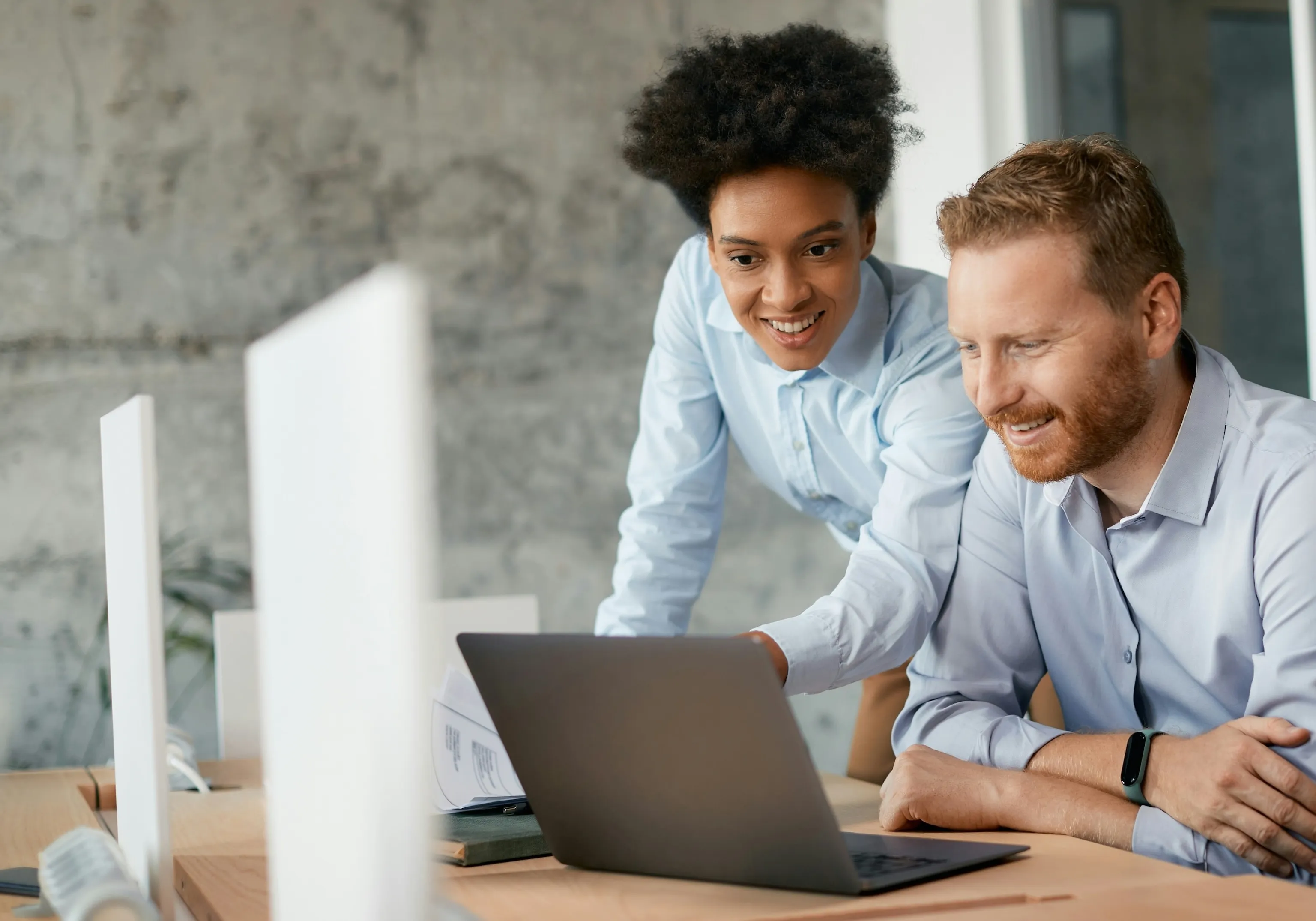 Two colleagues collaborating and smiling while looking at a laptop on a desk in an office.