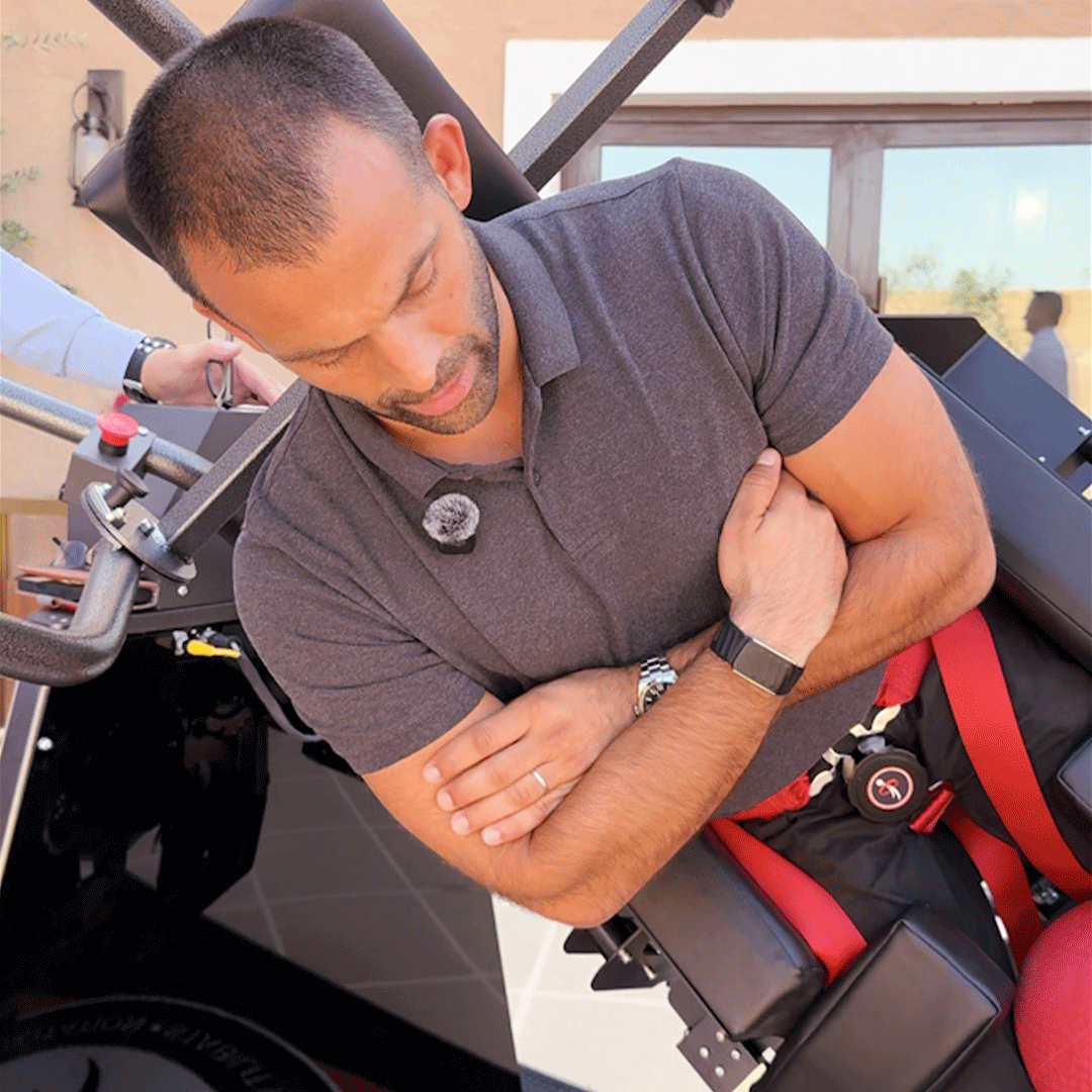 Man in a gray polo shirt using a black and red exercise machine, positioned sideways with arms crossed.