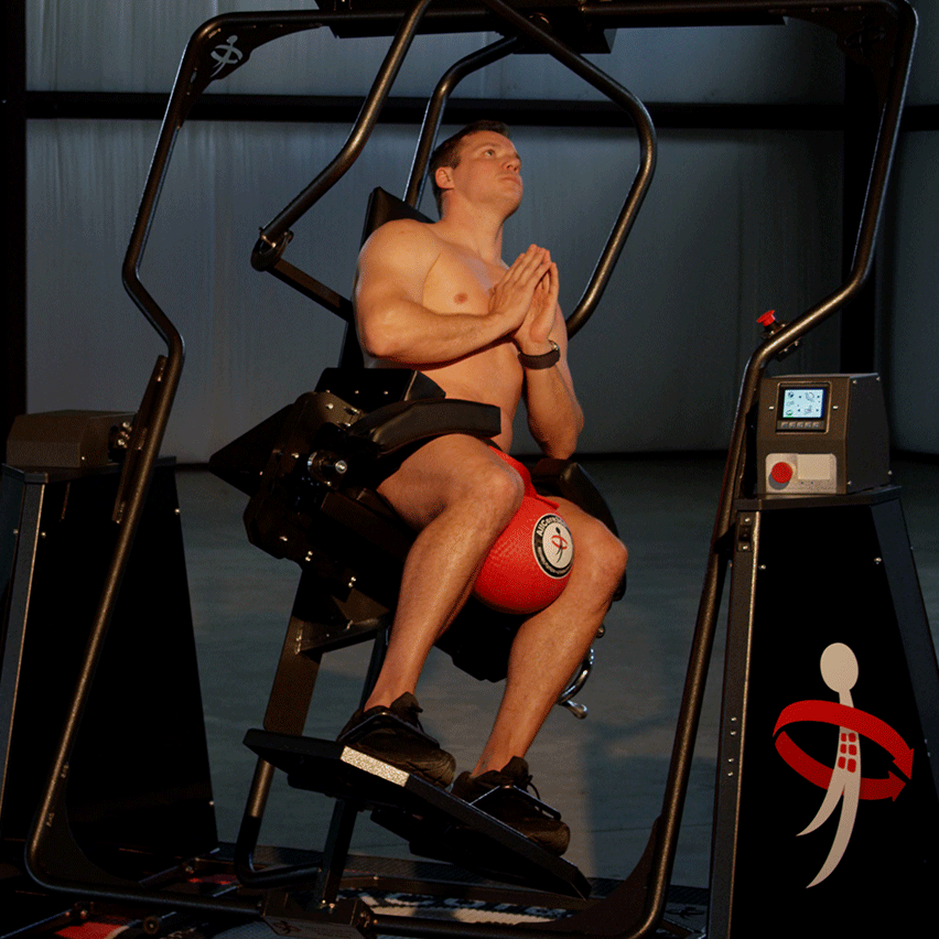 Shirtless man in black shorts exercising with a red medicine ball between his knees on a black fitness machine.