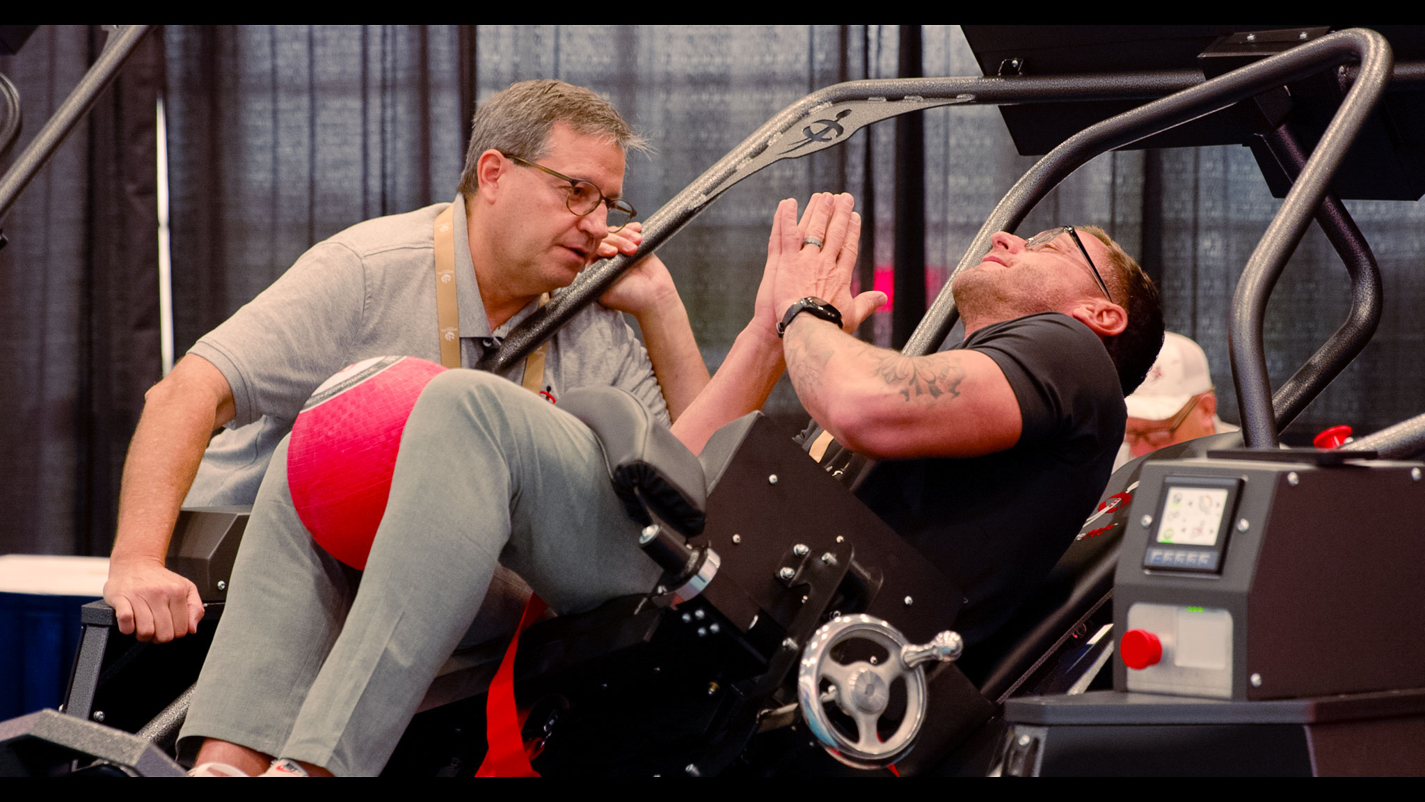 Man exercising on a leg press machine with a red ball between his legs while a trainer observes and gives a high five.
