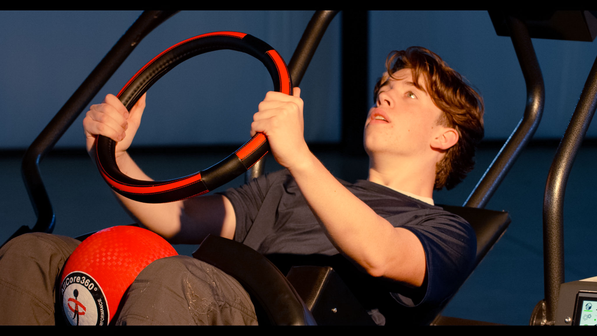 Young man gripping a black and red ring while performing an exercise on a fitness machine with a red AllCore360 performance ball on his lap.