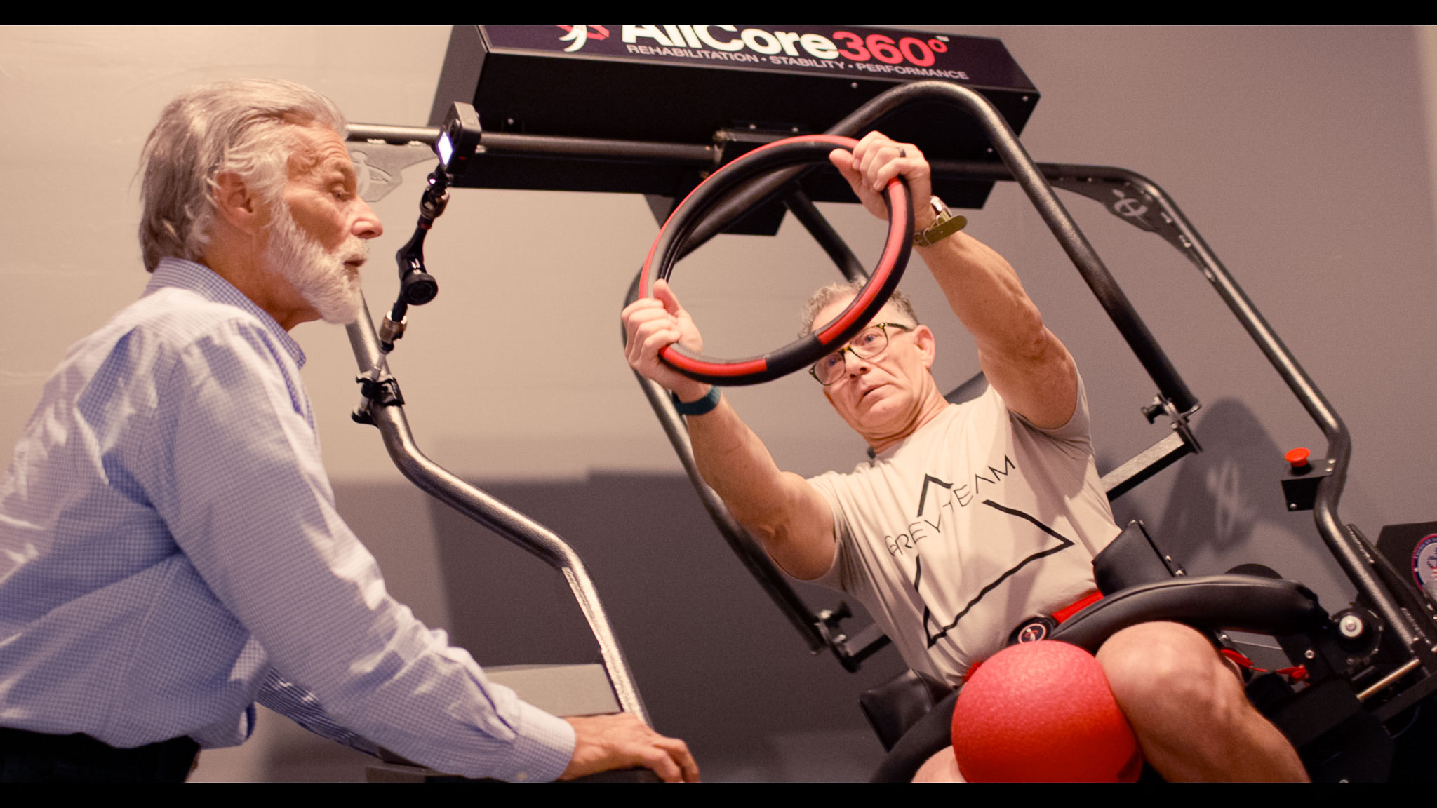 Older man in a Grey Team shirt exercising with a red ring on rehab equipment while another older man observes.