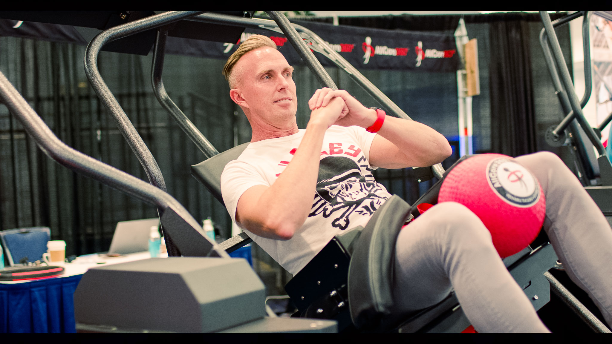 Man exercising on a leg press machine with a red exercise ball between his knees.
