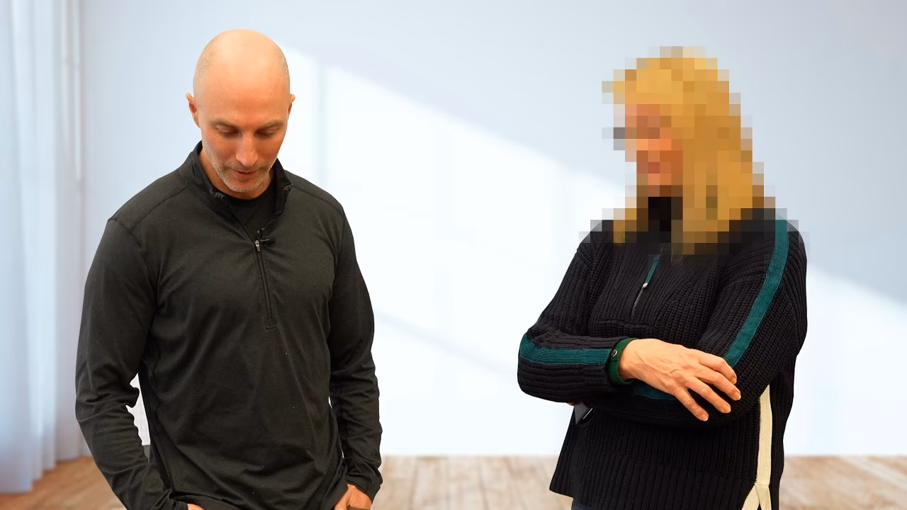 Bald man in a black zipped sweater and woman with pixelated face wearing a dark sweater with green and white stripes standing indoors.