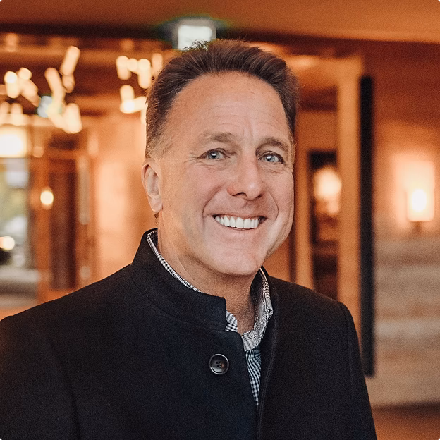 Smiling middle-aged man wearing a dark coat in a warmly lit indoor setting.