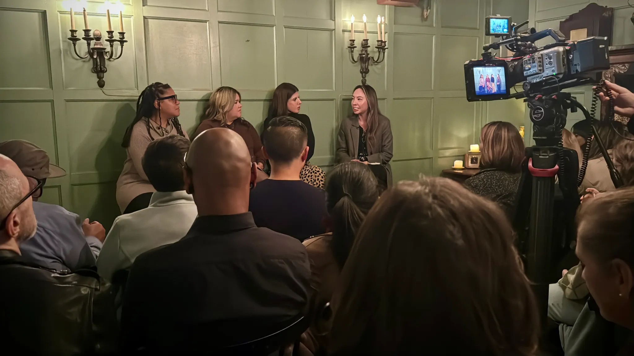 A panel discussion with four women seated in front of an audience, a video camera recording the event.