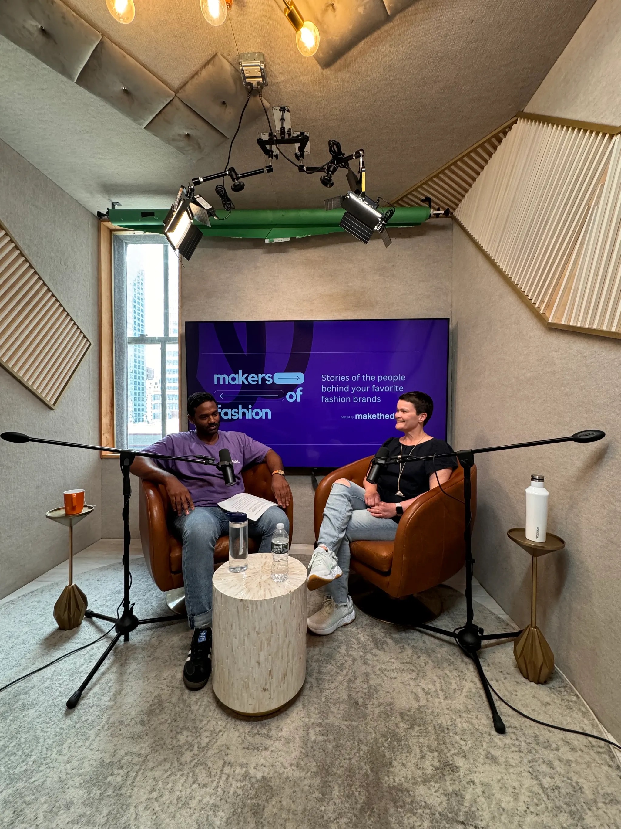 Two people sitting in brown leather chairs with microphones discussing in a podcast studio with a screen behind them reading 'makers of fashion.'