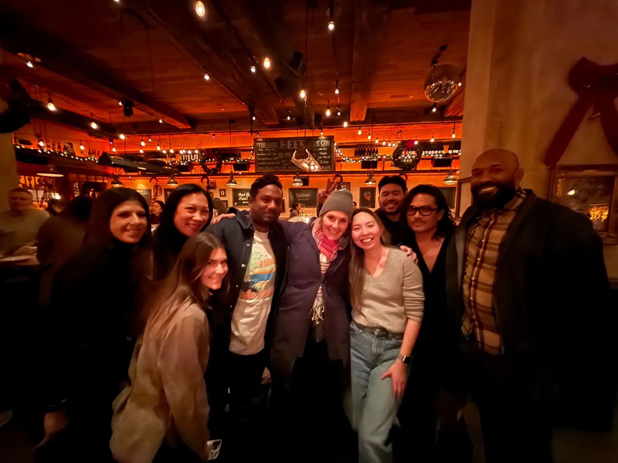 Group of eight people smiling and posing together in a warmly lit bar or restaurant with festive string lights and wood ceiling.