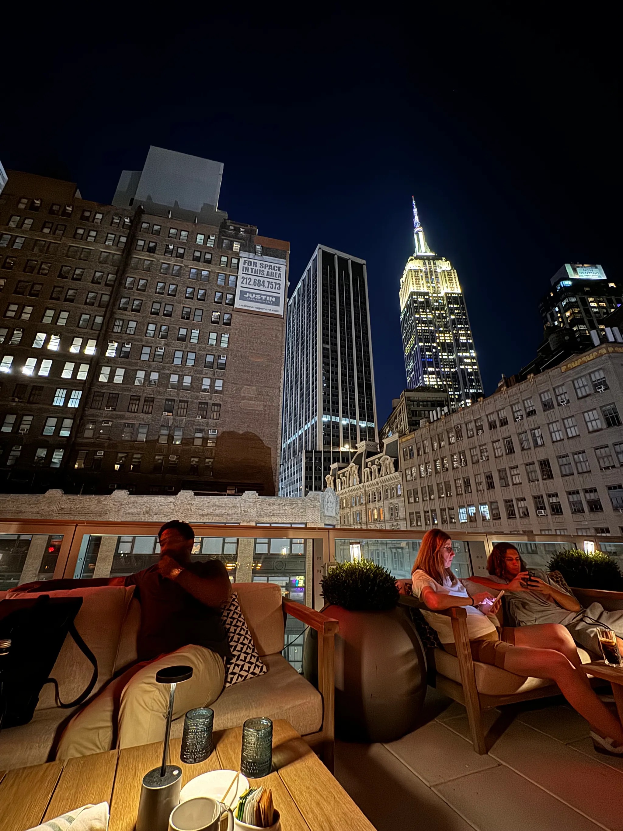People sitting on a rooftop patio at night with the illuminated Empire State Building and surrounding city skyscrapers in the background.