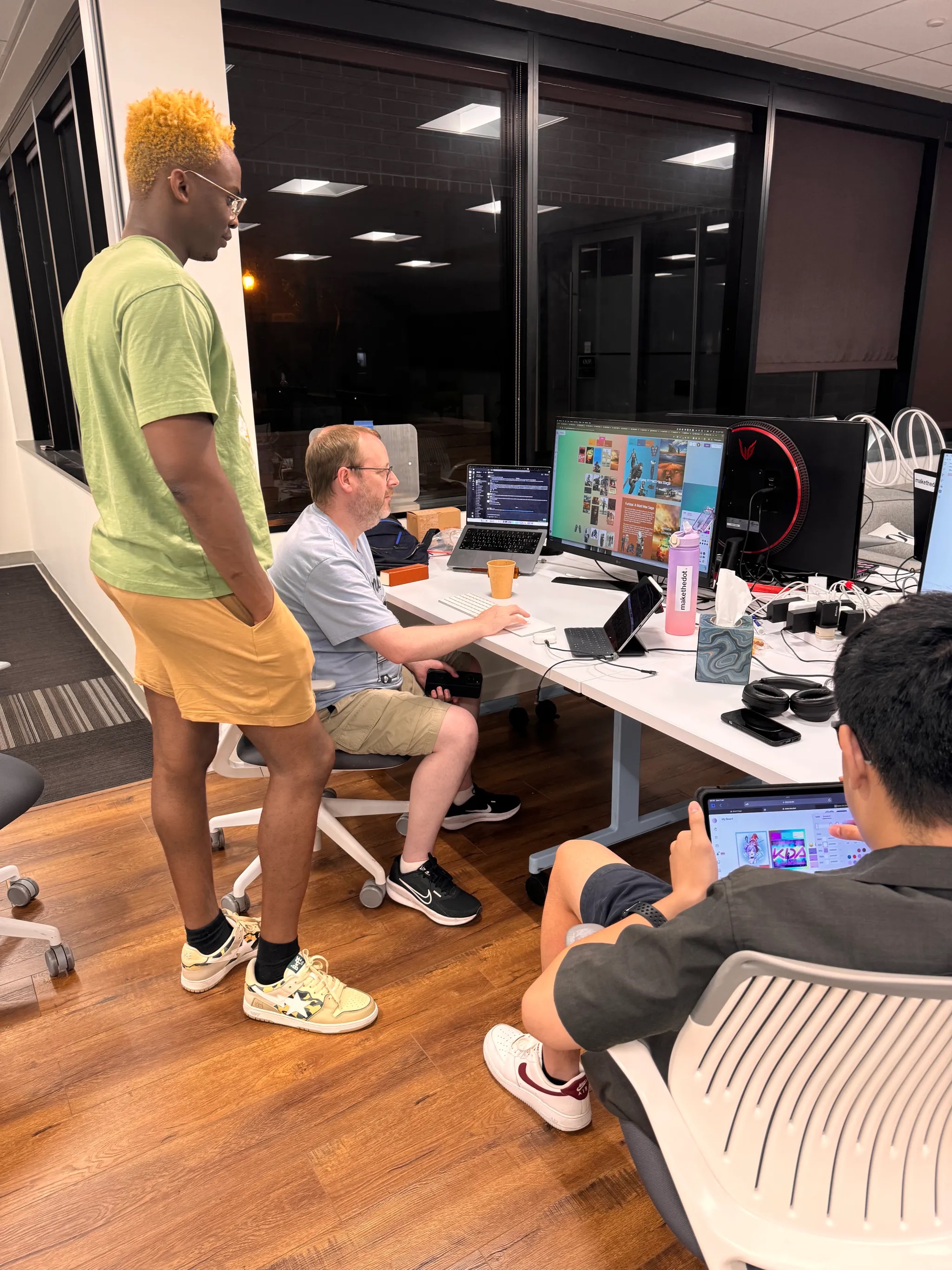 Three men in casual clothing collaborating at a desk with multiple computer screens and devices in an office setting.