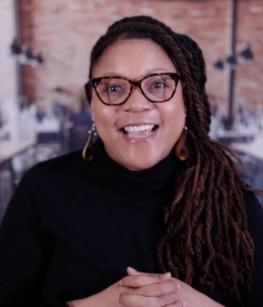 Smiling woman with glasses and long braided hair wearing a black turtleneck in an indoor setting.