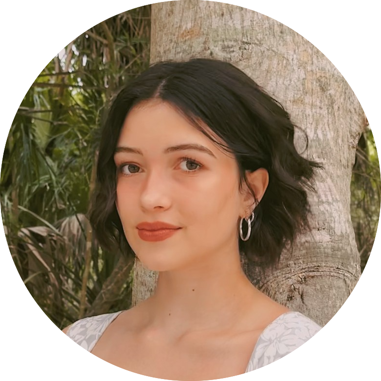 Young woman with short dark hair and silver hoop earrings, standing in front of a tree with greenery in the background.