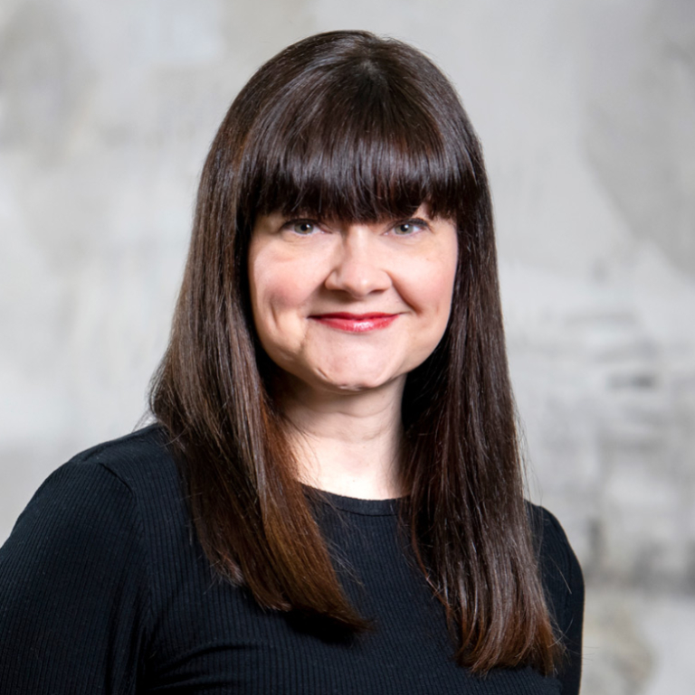 Smiling woman with straight dark brown hair and bangs wearing a black top against a light blurred background.
