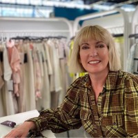 Smiling woman with short blonde hair wearing a brown plaid shirt sitting in front of a clothing rack with neutral-colored garments.