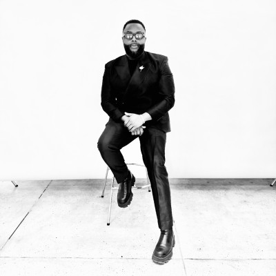 A man dressed in a black suit and glasses sits on a stool against a plain white background.