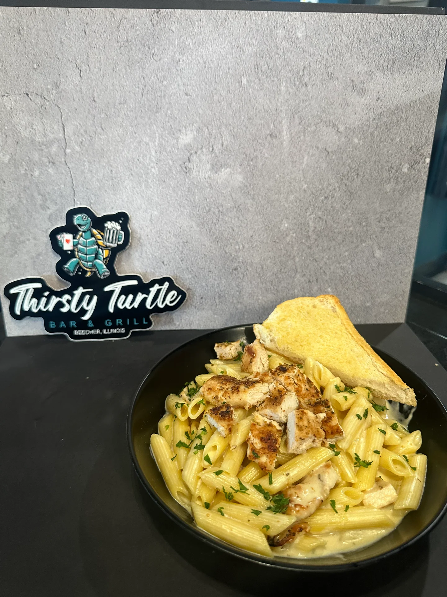 Black bowl of penne pasta with grilled chicken, cream sauce, sprinkled with parsley, and a slice of toasted bread on the side.