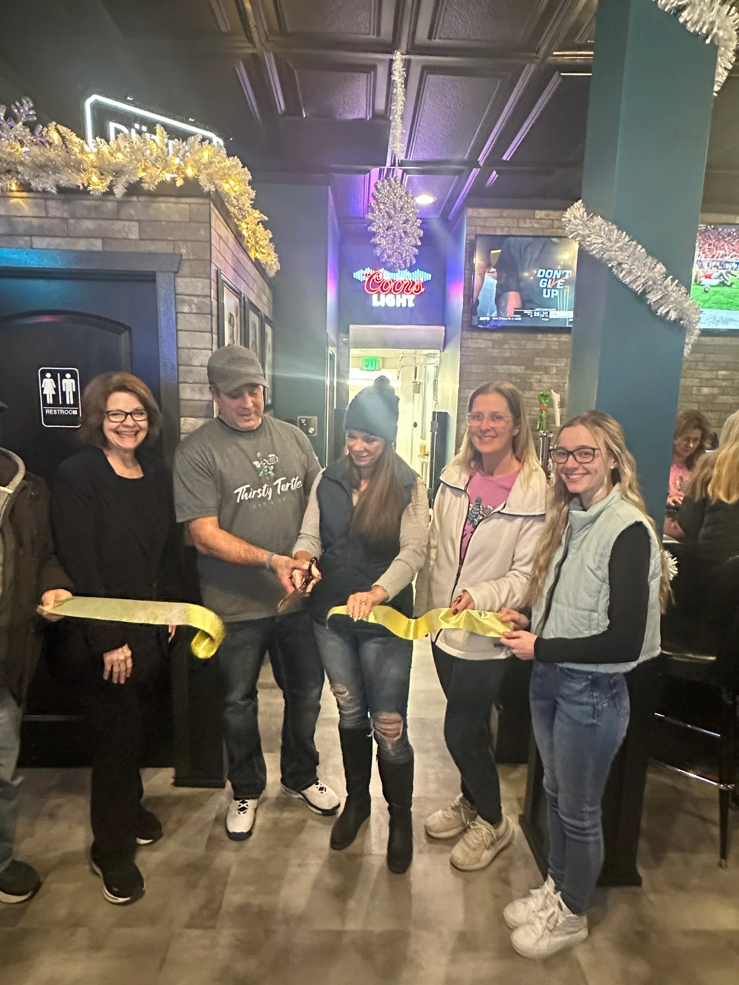 Five people standing indoors cutting a yellow ribbon during an event, with festive silver garlands and a Coors Light sign in the background.