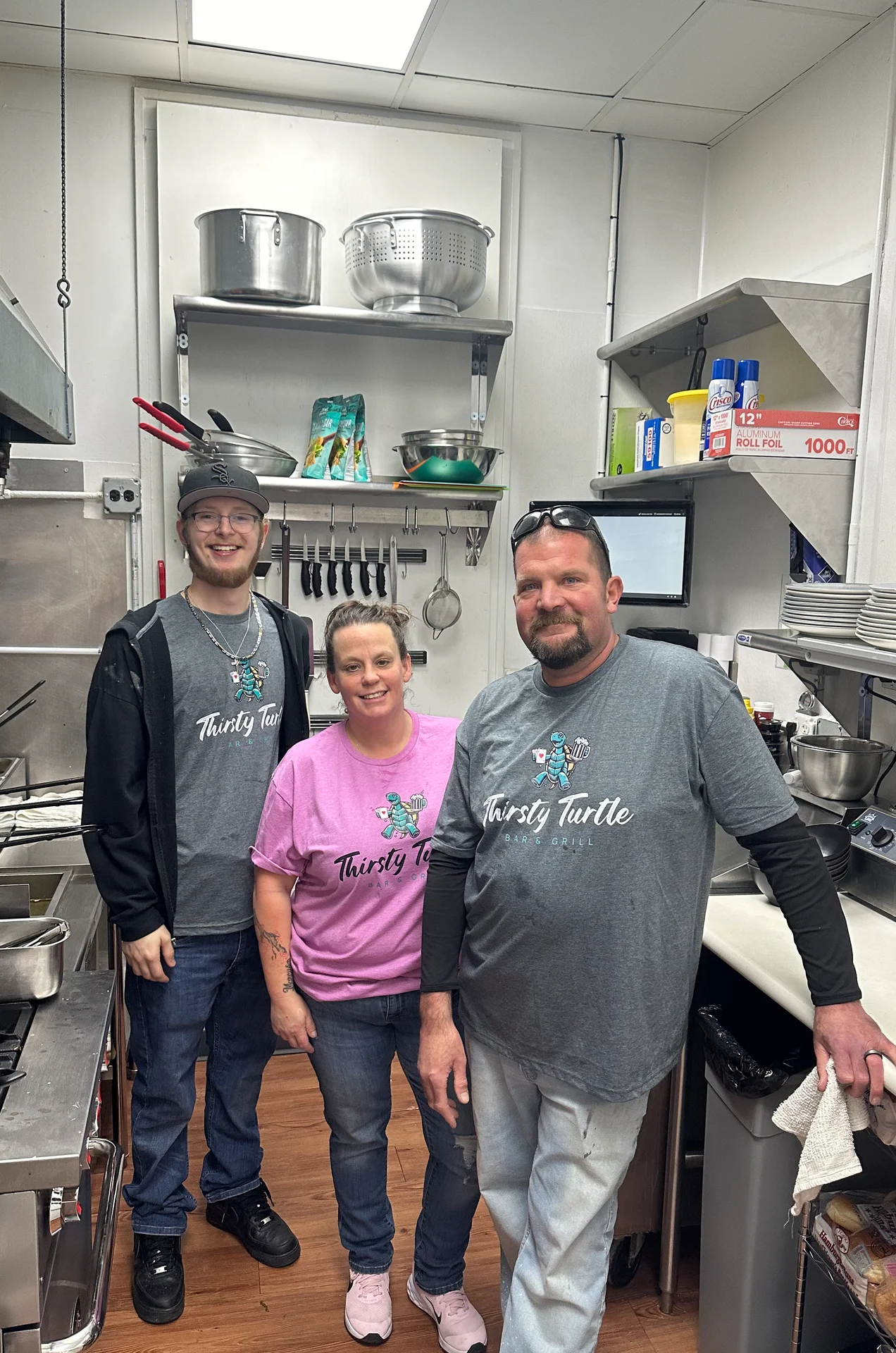 Three people standing and smiling in a commercial kitchen wearing Thirsty Turtle Bar & Grill shirts.