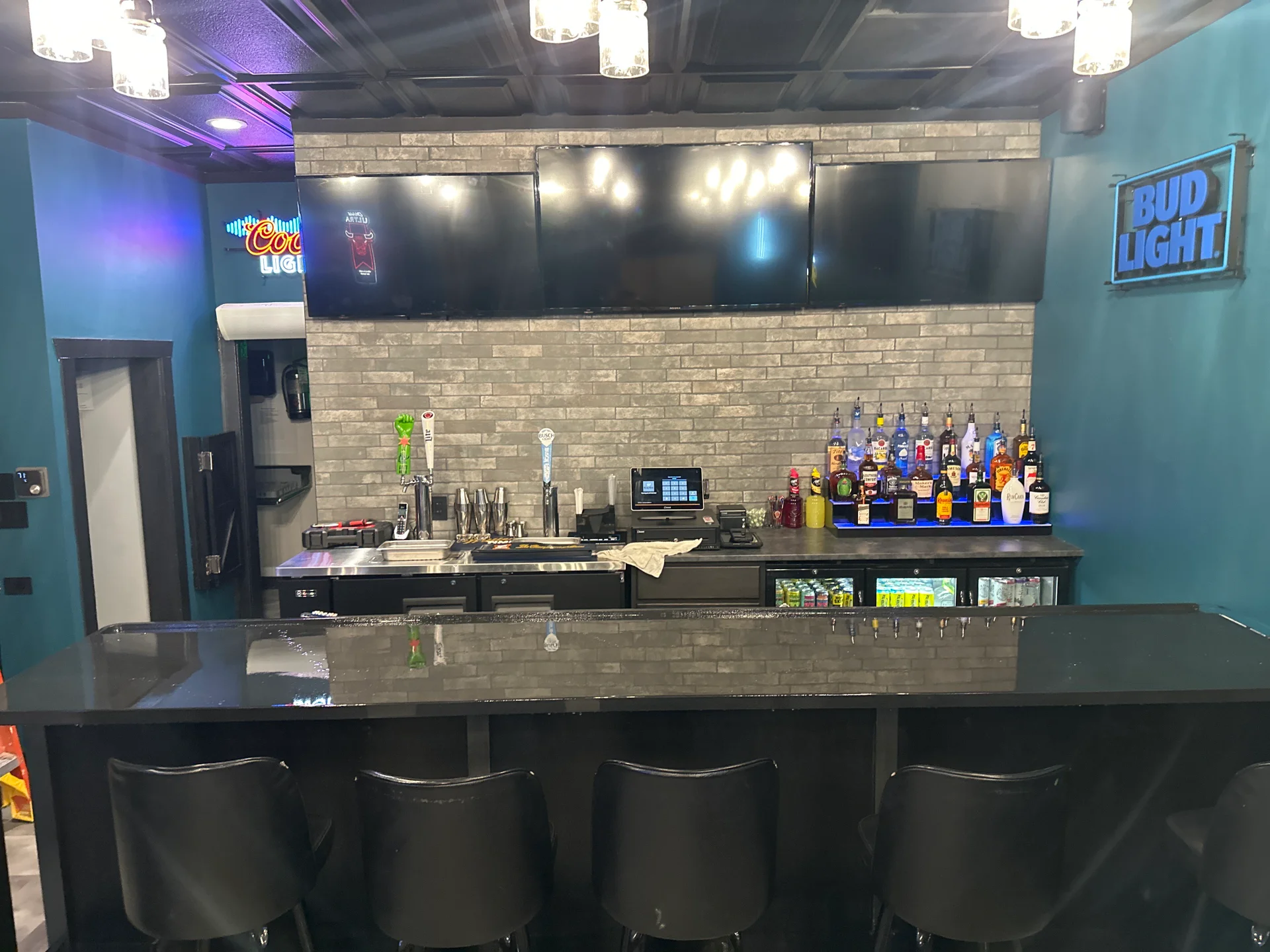 Modern bar with black counter, black stools, multiple liquor bottles on display, three large TVs mounted on a gray brick wall, and neon Bud Light and Coors Light signs.