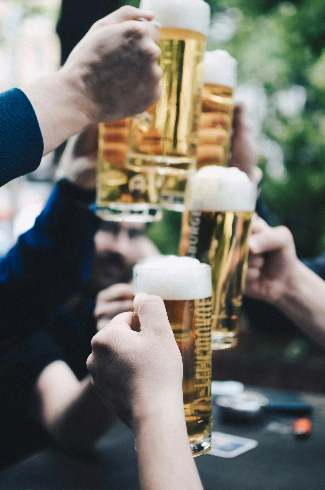 Several people clinking tall glasses of beer with foam in an outdoor setting.