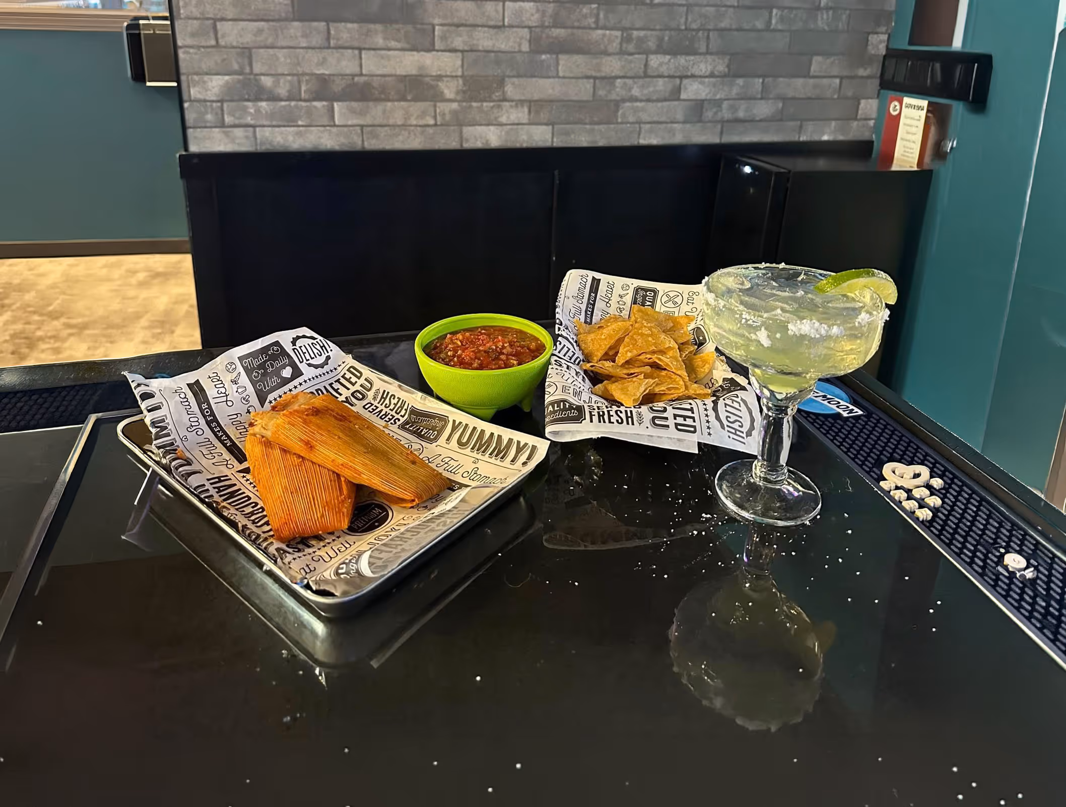Tray with two tamales, green bowl of salsa, basket of tortilla chips, and a salted margarita glass with lime wedge on a black reflective table.