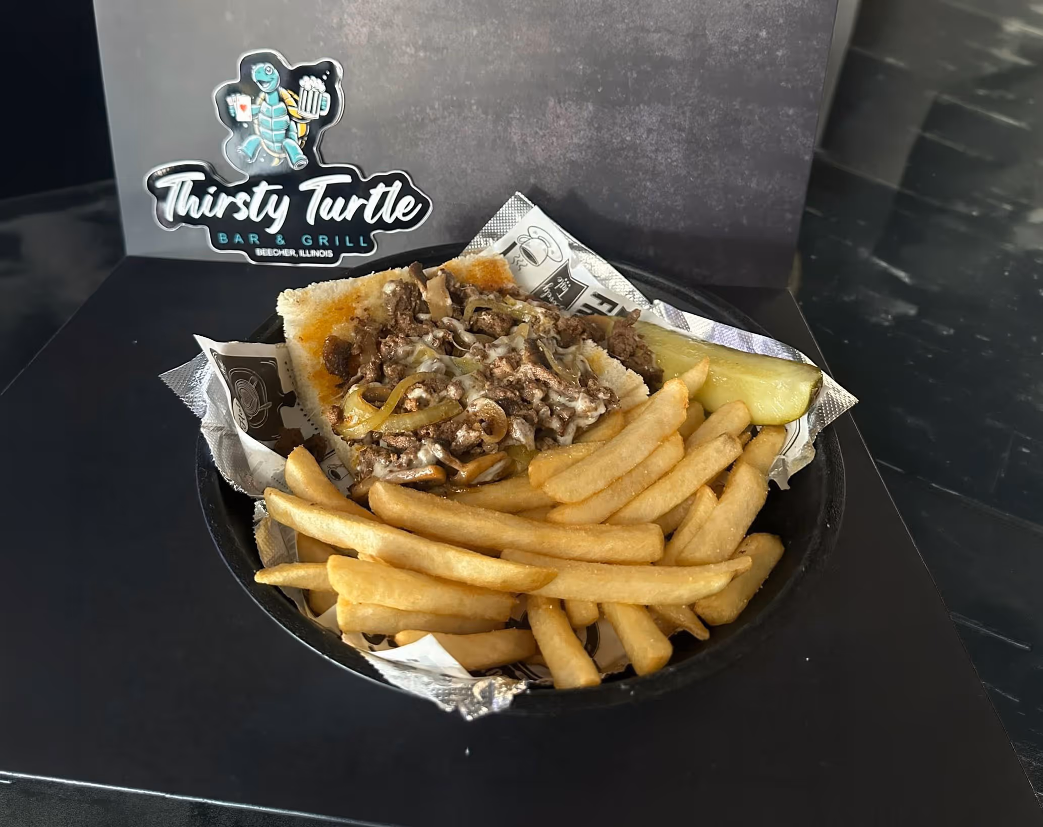 Sandwich with ground beef, melted cheese, and onions served with French fries and a pickle slice in a black tray.