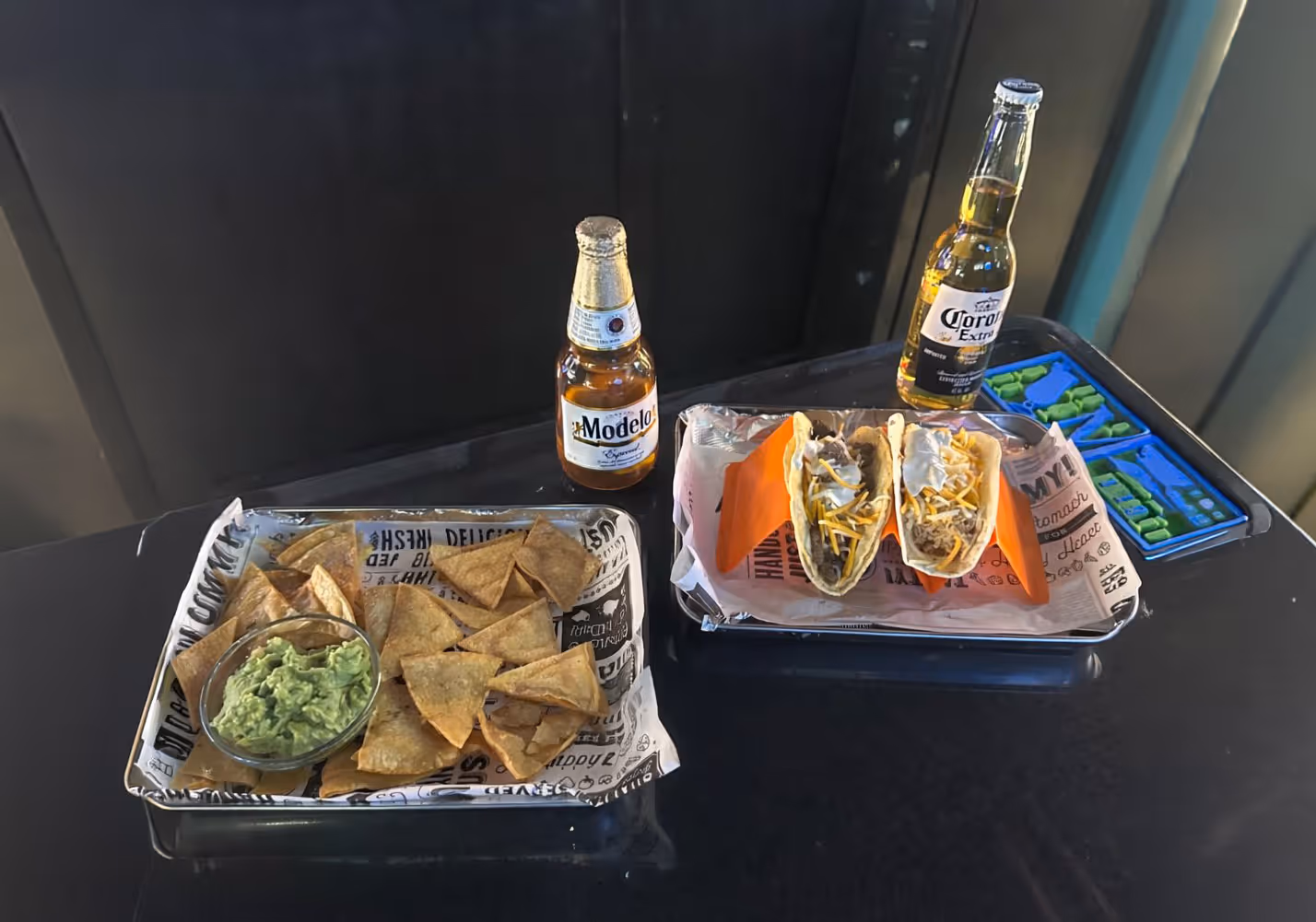 Two trays on a black table; one has tortilla chips with guacamole and a Modelo beer, the other has two tacos with shredded cheese and sour cream alongside a Corona Extra beer.