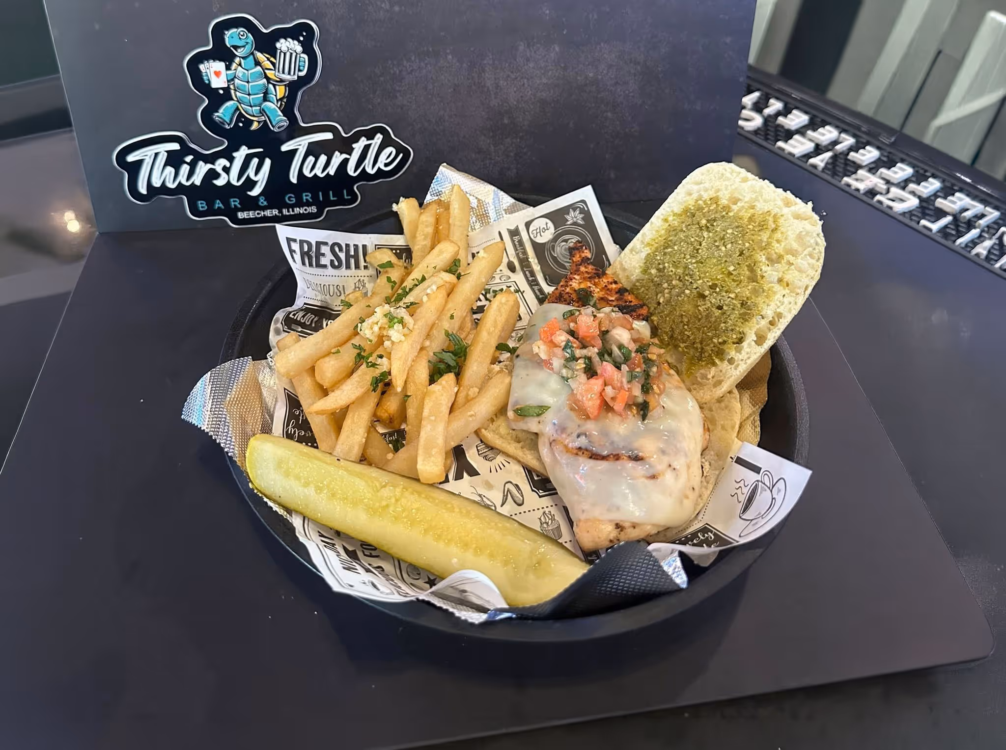 Open chicken sandwich with melted cheese, pico de gallo, pesto spread, fries topped with parsley and garlic, and a pickle spear in a black basket lined with newspaper-style paper.
