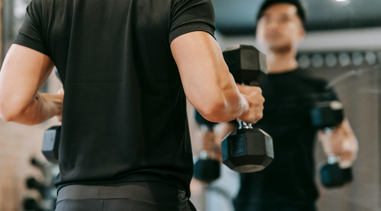 A person in a gym is seen lifting dumbbells, wearing a black outfit, and focusing intensely in a mirror. The atmosphere is energetic and determined.