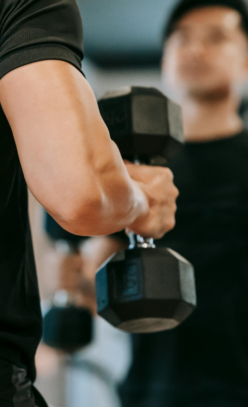 A person in a gym is seen lifting dumbbells, wearing a black outfit, and focusing intensely in a mirror. The atmosphere is energetic and determined.