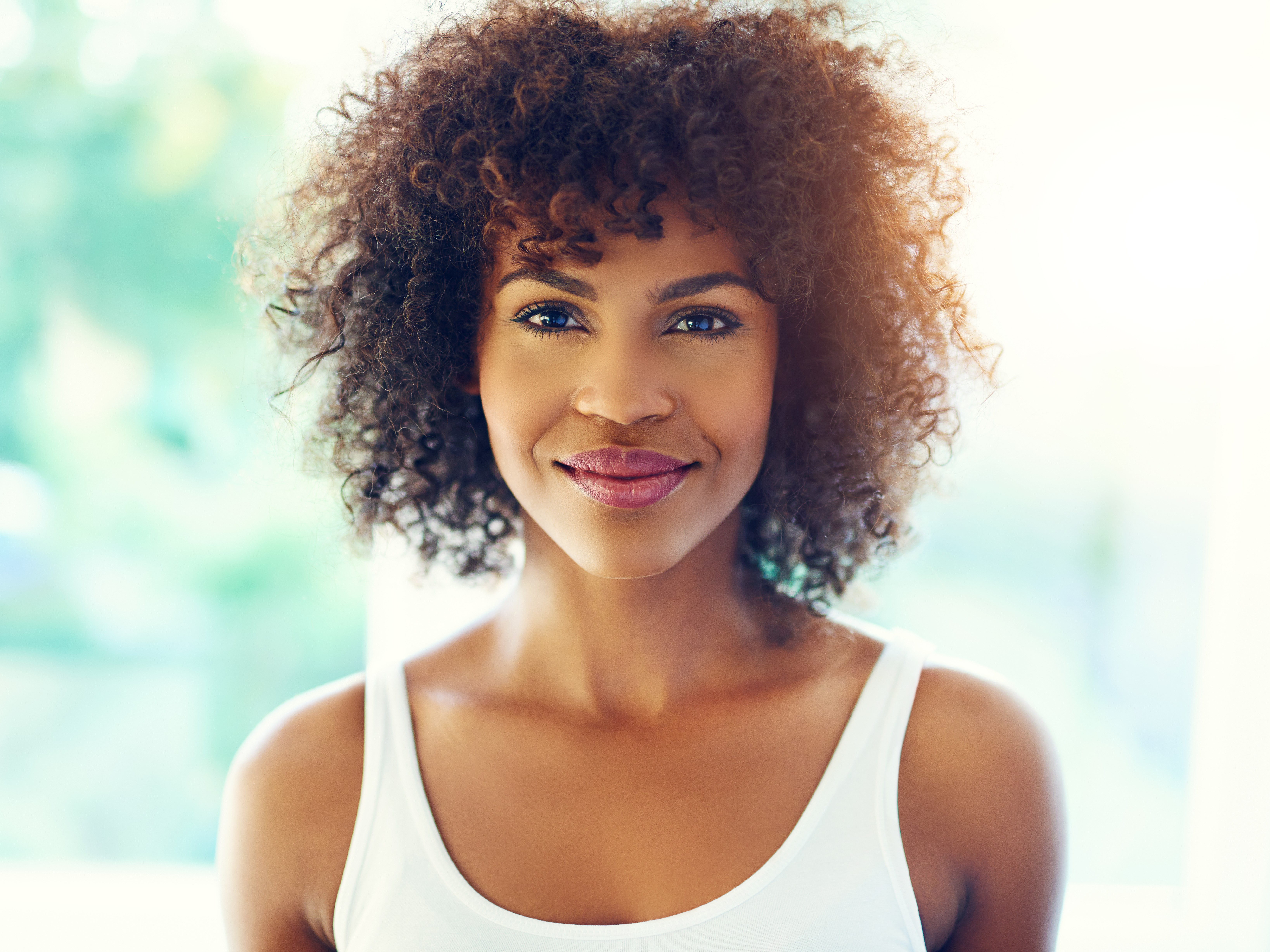 Smiling woman with curly hair wearing a white tank top against a bright, blurred background.