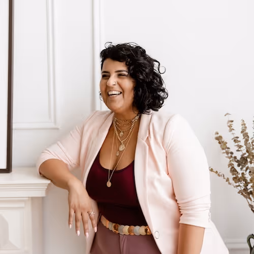 Smiling woman with curly black hair wearing layered gold necklaces, maroon top, light pink blazer, and mauve pants, leaning on a white mantle.