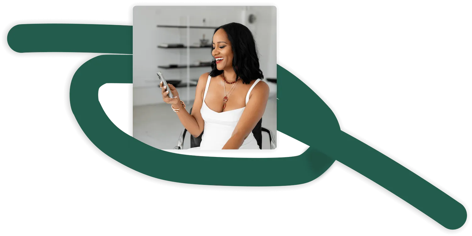 Smiling woman in white dress sitting on a chair looking at her smartphone indoors.