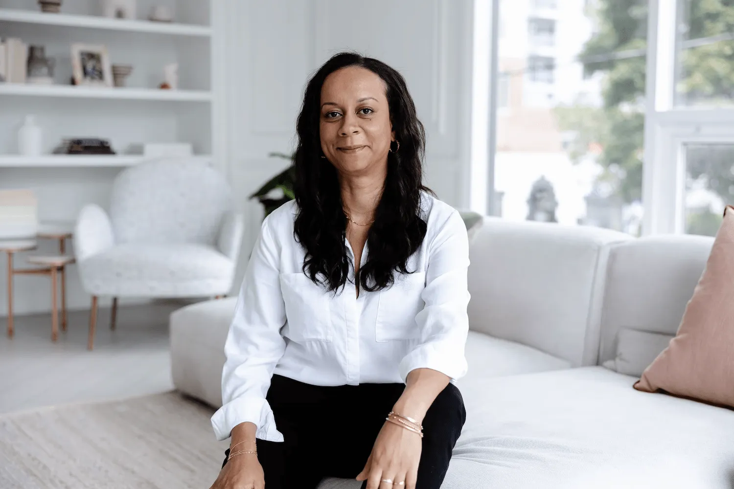 Woman with long dark hair wearing a white shirt and black pants sitting on a light-colored sofa in a bright living room.