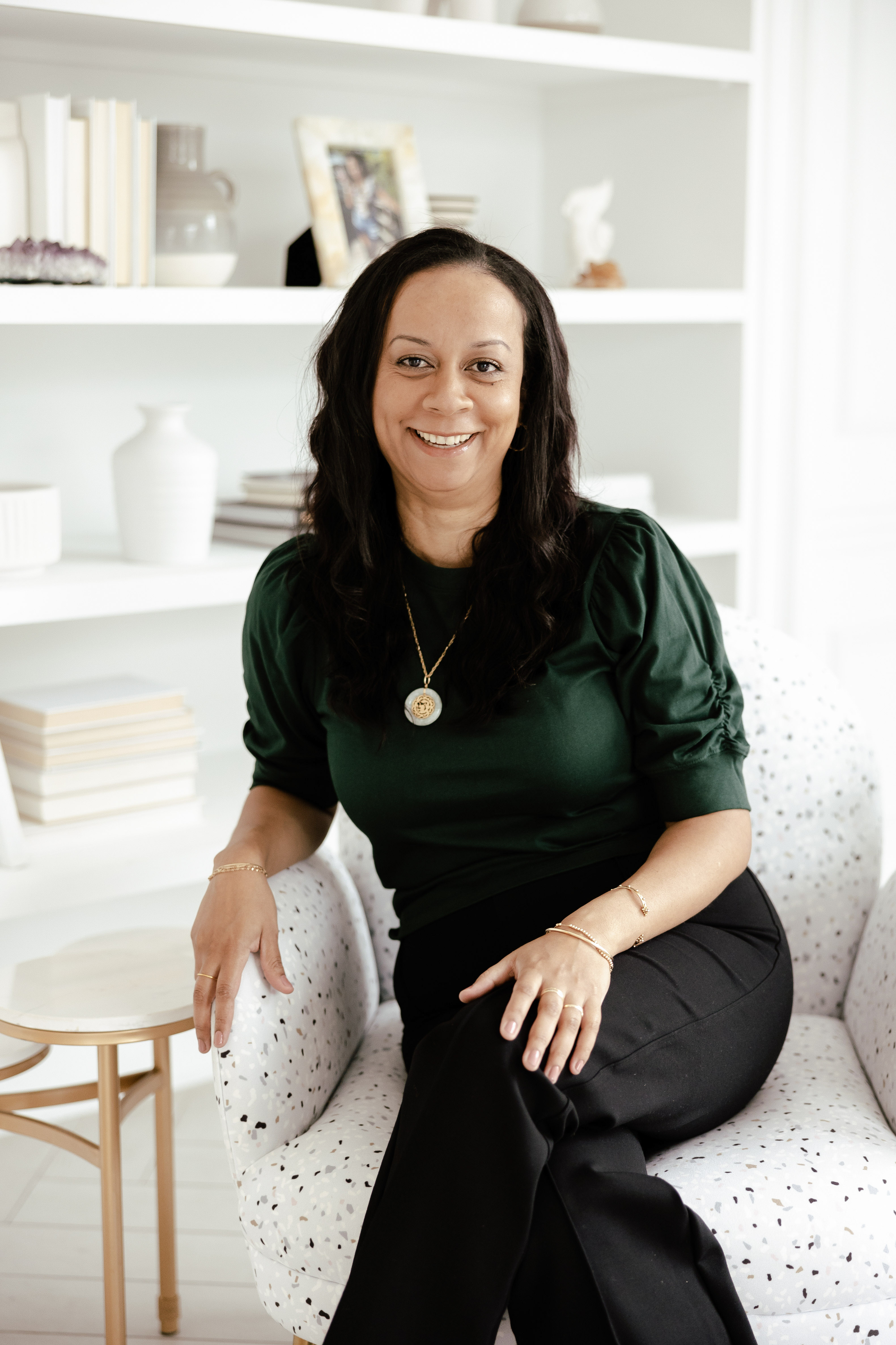 Smiling woman with long hair wearing green top sitting cross-legged on a patterned armchair indoors.