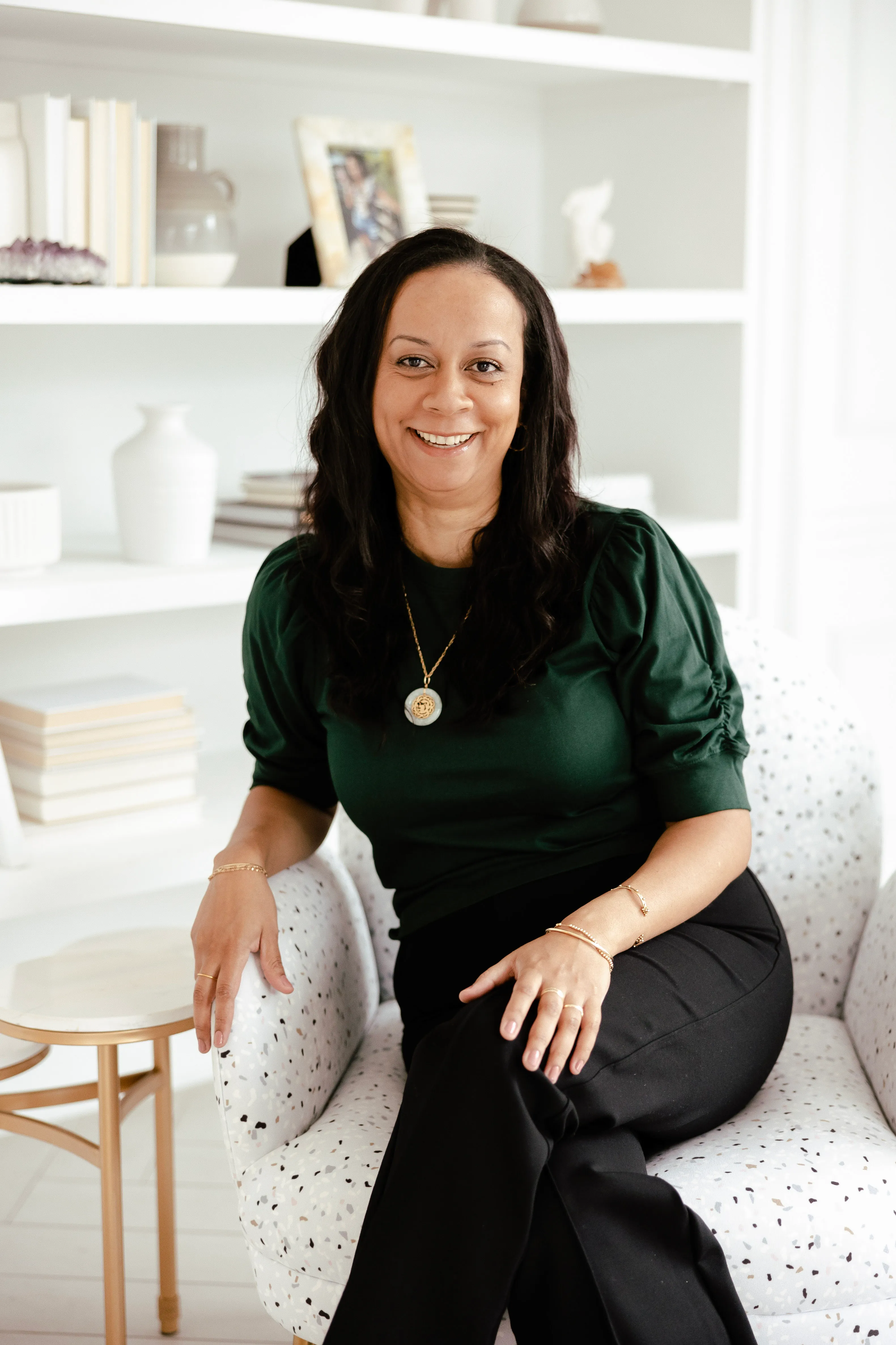 Smiling woman with long hair wearing green top sitting cross-legged on a patterned armchair indoors.