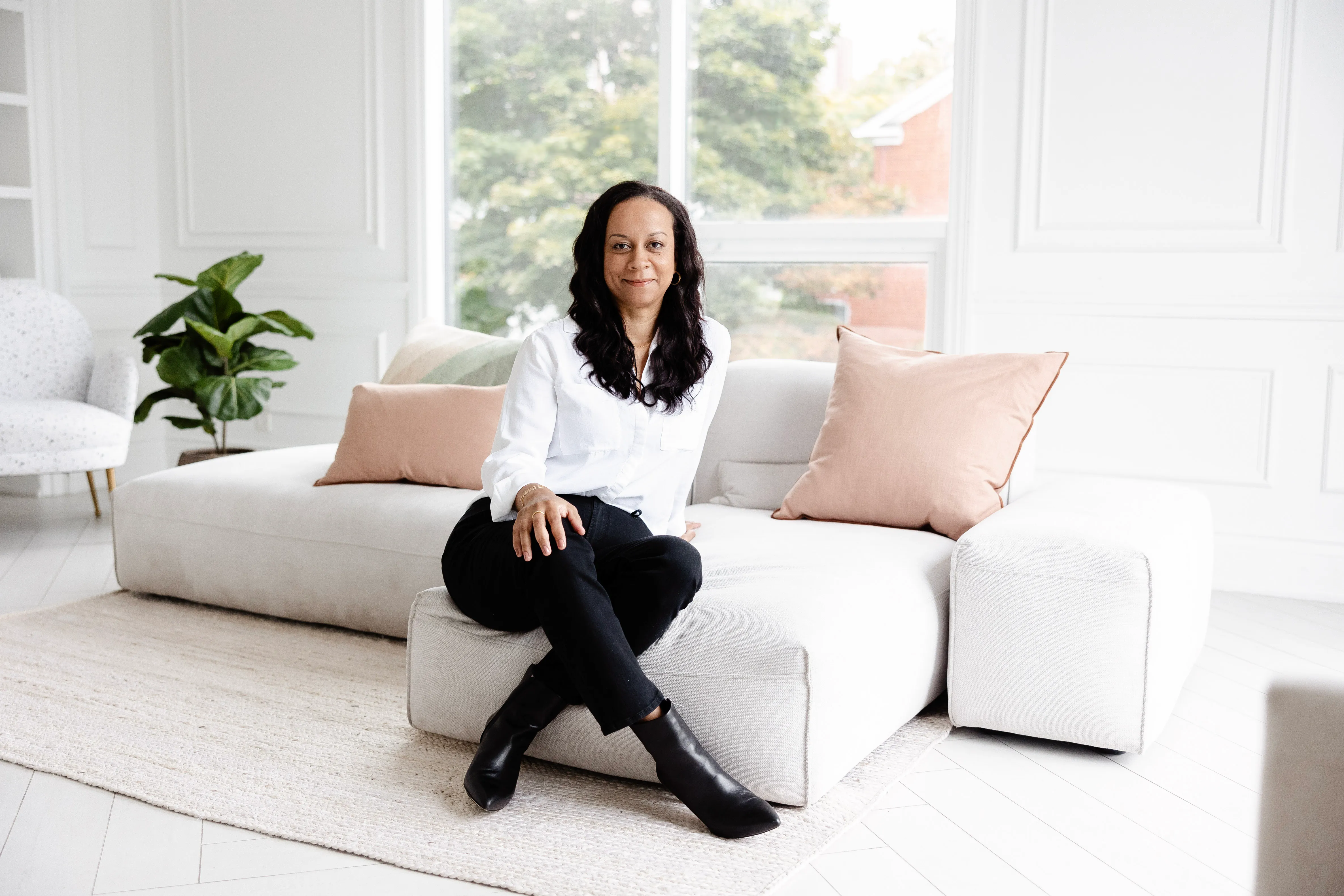 Woman with long dark hair sitting on a light sofa in a bright room with large window and pink pillows.