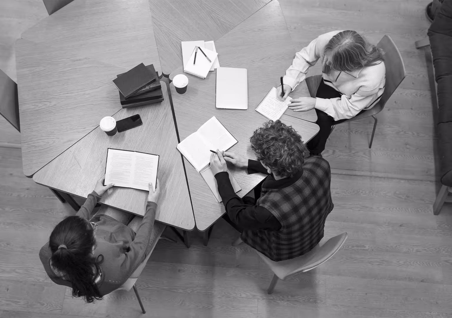 Tres estudiantes sentados alrededor de una mesa triangular tomando notas y leyendo libros.