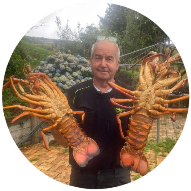 Man outdoors holding two large lobsters with a garden and hydrangea flowers in the background.