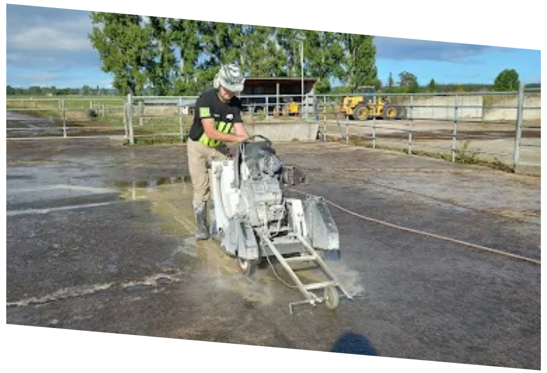 Person operating a concrete saw machine on an outdoor paved surface near metal fencing.