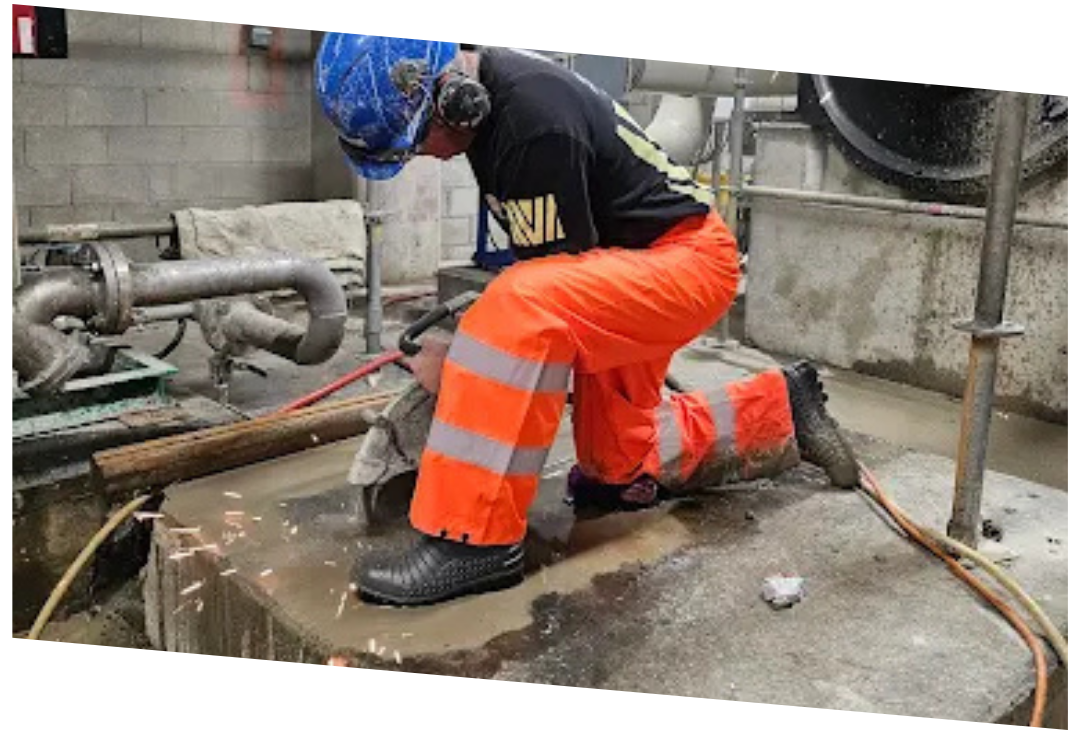 Worker in orange reflective pants and blue helmet cutting concrete with a power saw indoors.