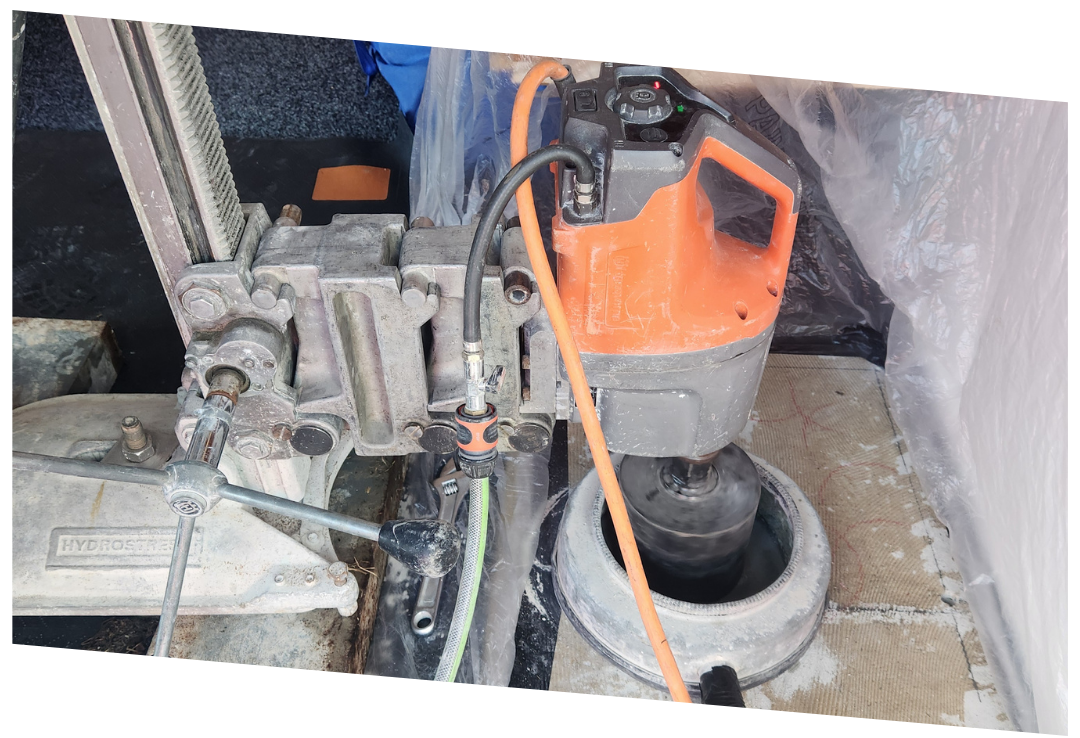 Close-up of an orange and gray core drilling machine with hoses attached, drilling into a concrete surface.