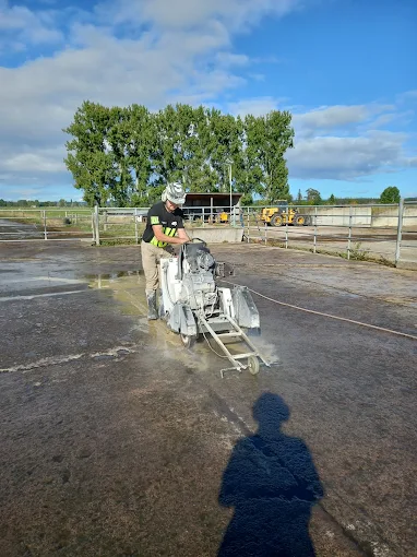 Person using a machine to cut concrete on an outdoor construction site with trees and equipment in the background.