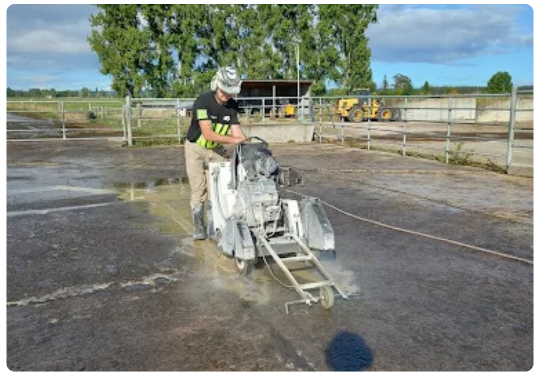 Person operating a concrete cutting machine on a large outdoor concrete surface near metal railings.