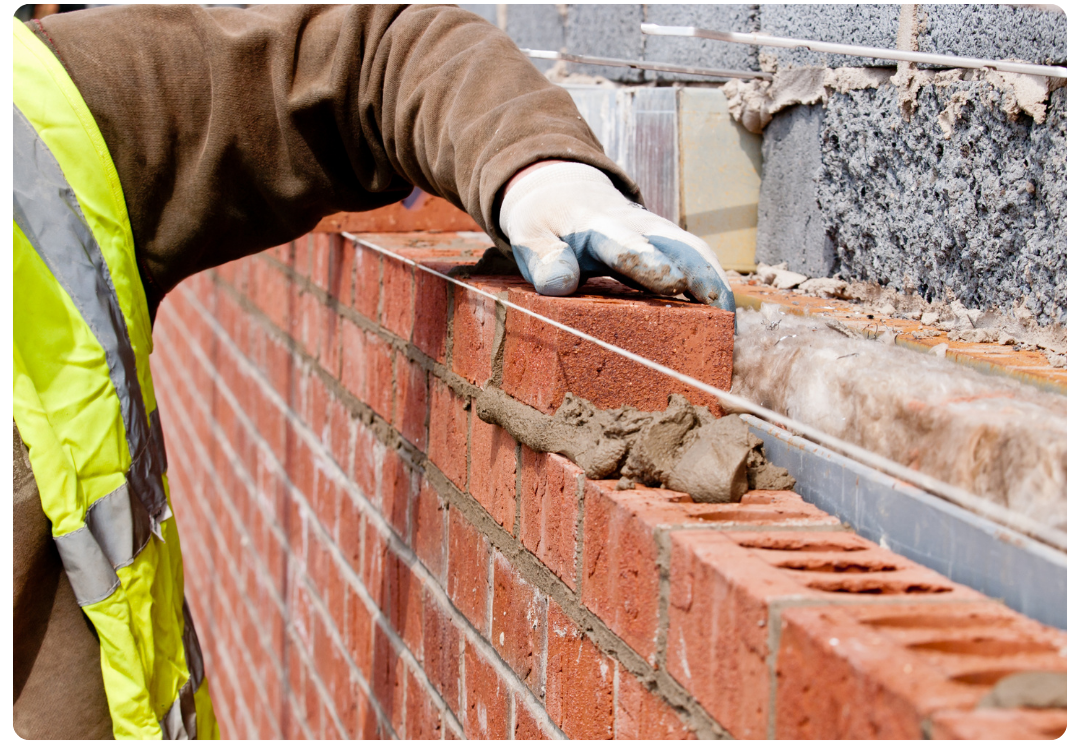 Construction worker wearing gloves and a safety vest placing a red brick on a wall with mortar.