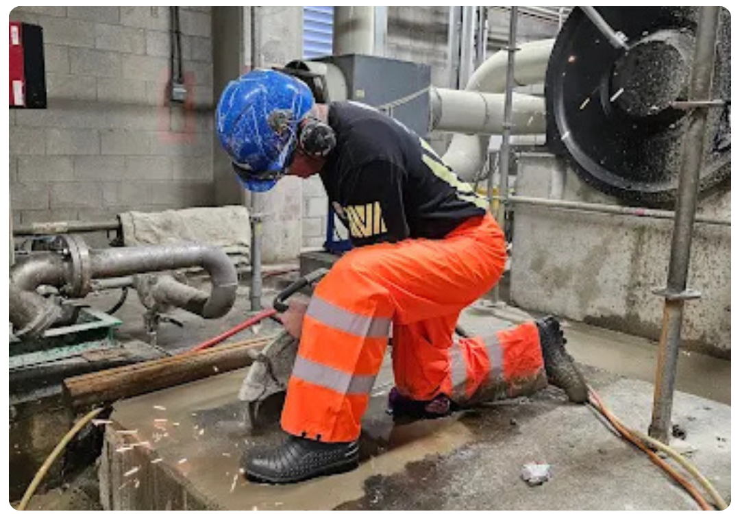 Worker in reflective orange pants and blue helmet cutting metal with sparks in an industrial setting.