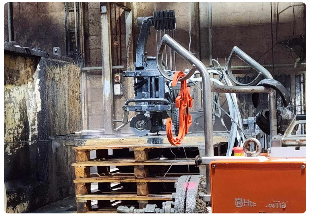 Industrial machine with orange hooks hanging, wooden pallets stacked, and worn concrete walls in a factory setting.