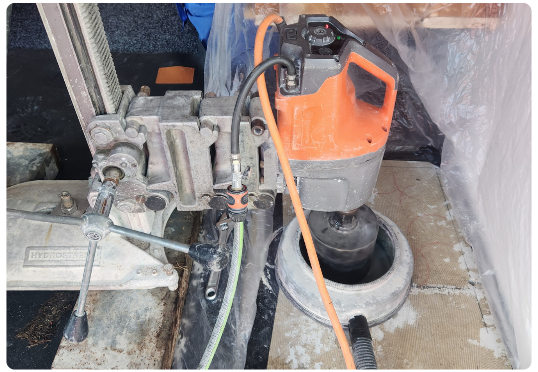 Core drilling machine with orange motor drilling into concrete, secured on a metal base.