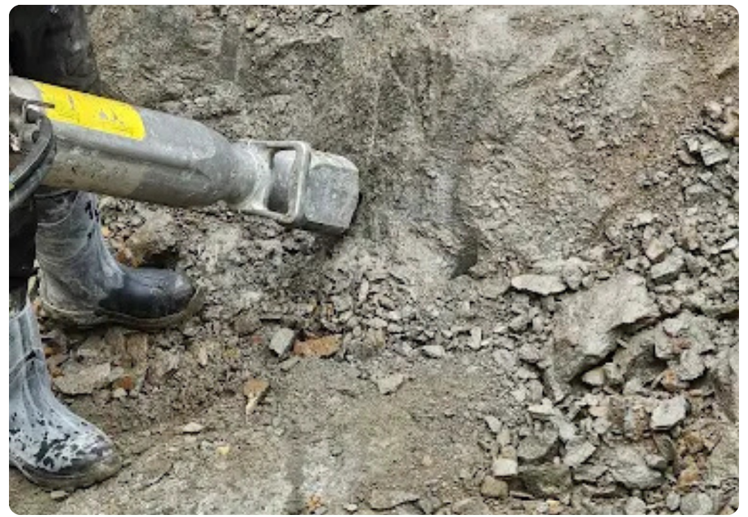 Concrete breaking tool operated by a person wearing black rubber boots on rubble ground.